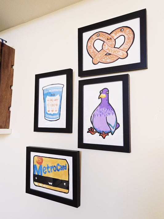 Four framed illustrations by The Carrot Press on a wall, including a pretzel, a pigeon, a MetroCard, and a blue coffee cup.