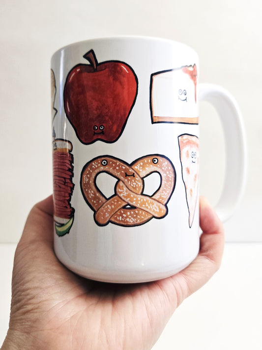 White mug with illustrations of an NYC apple, pretzel, and nyc cheesecake held by a hand against a white background