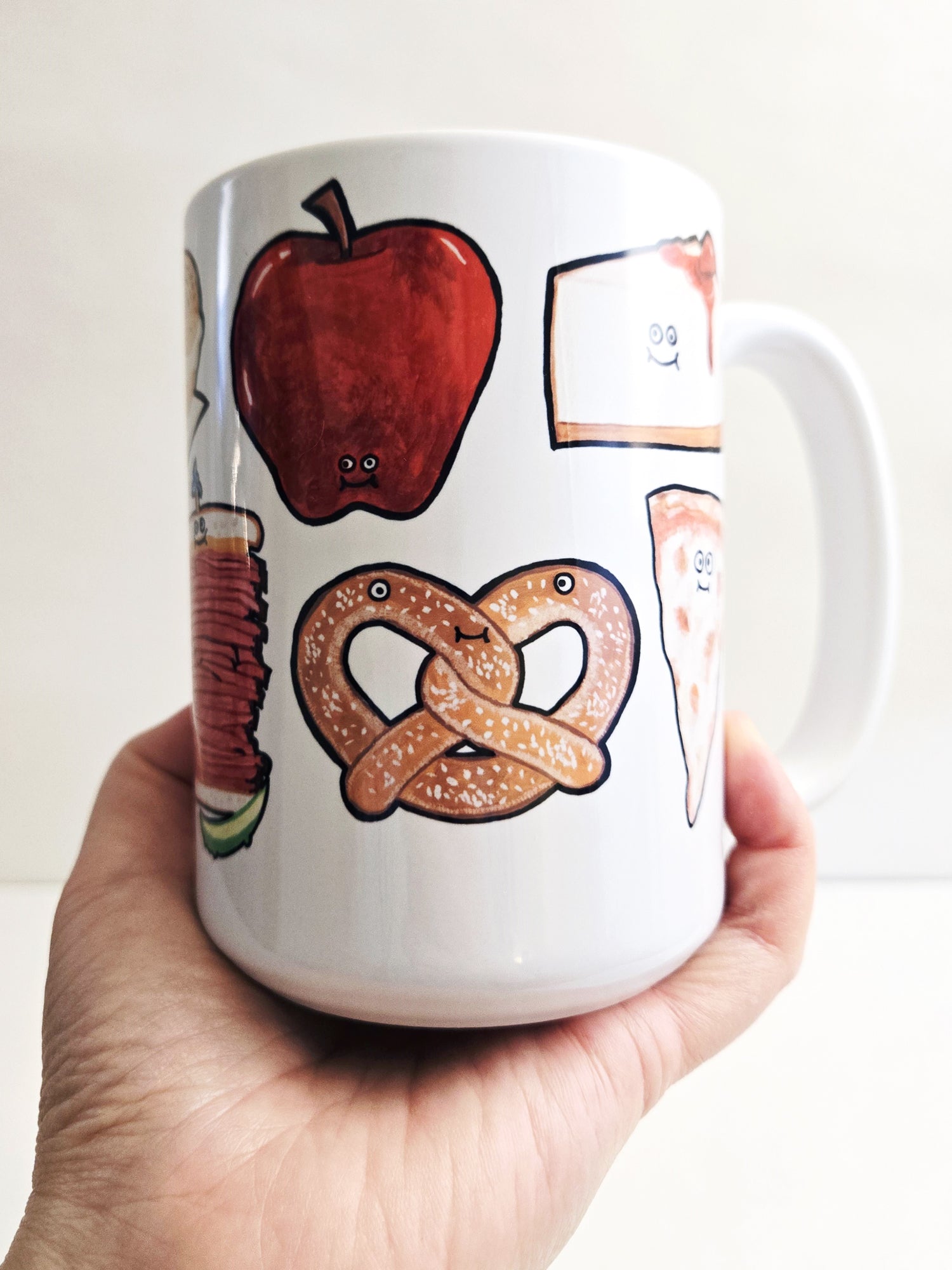 White mug with illustrations of an NYC apple, pretzel, and nyc cheesecake held by a hand against a white background