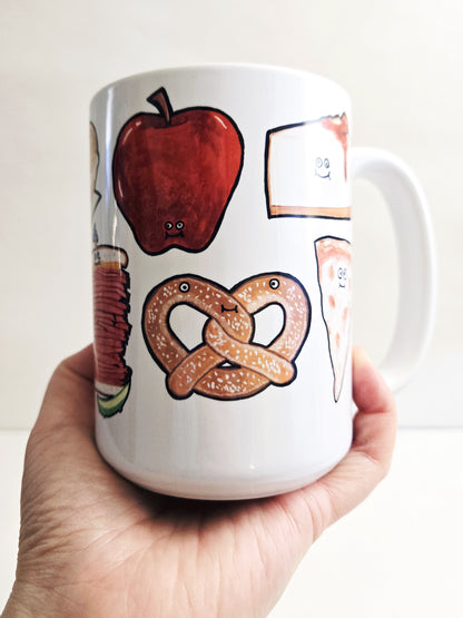 White mug with illustrations of an NYC apple, pretzel, and nyc cheesecake held by a hand against a white background