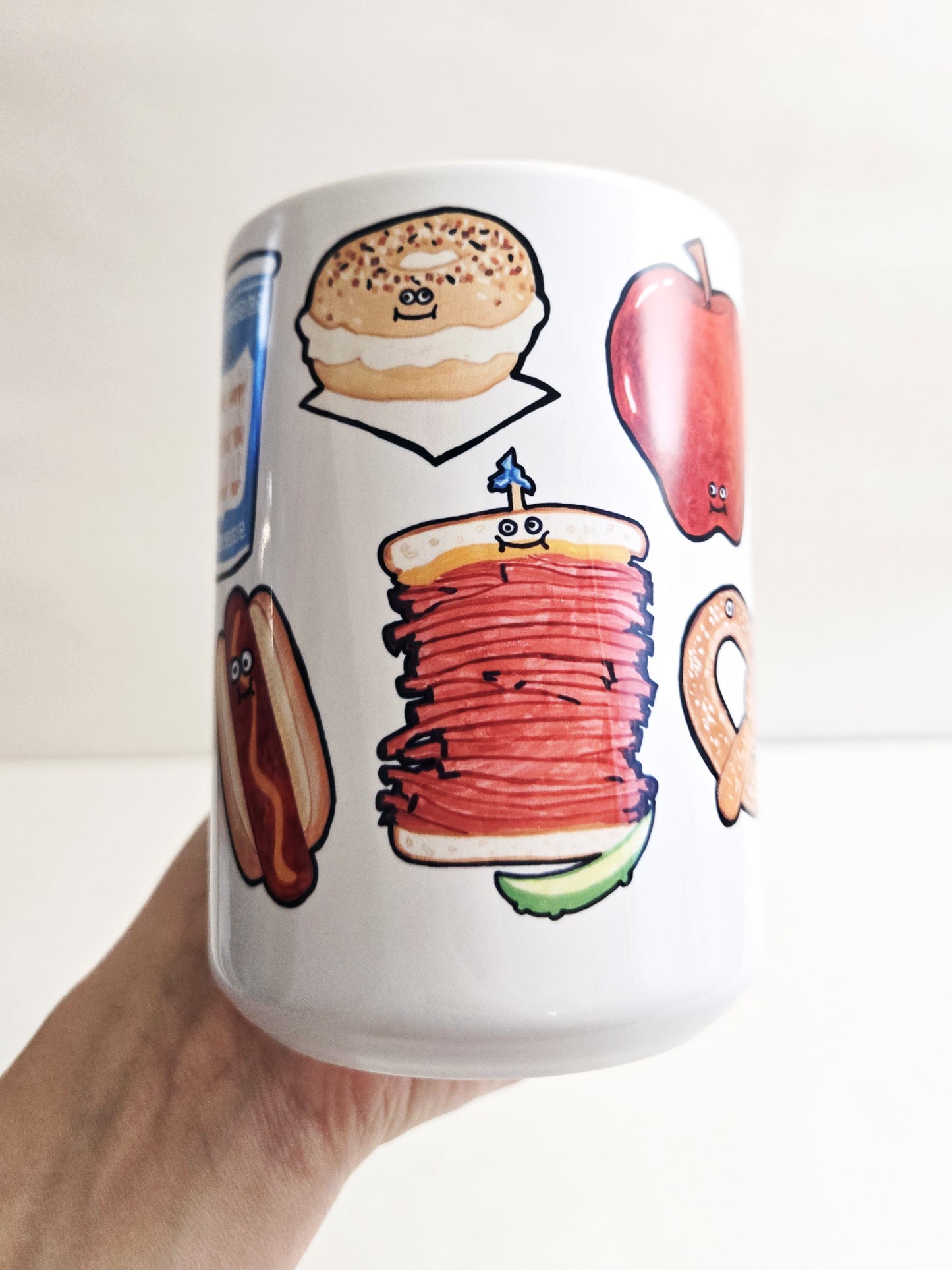 Mug with New York City 
food-themed illustrations held by a hand against a white background