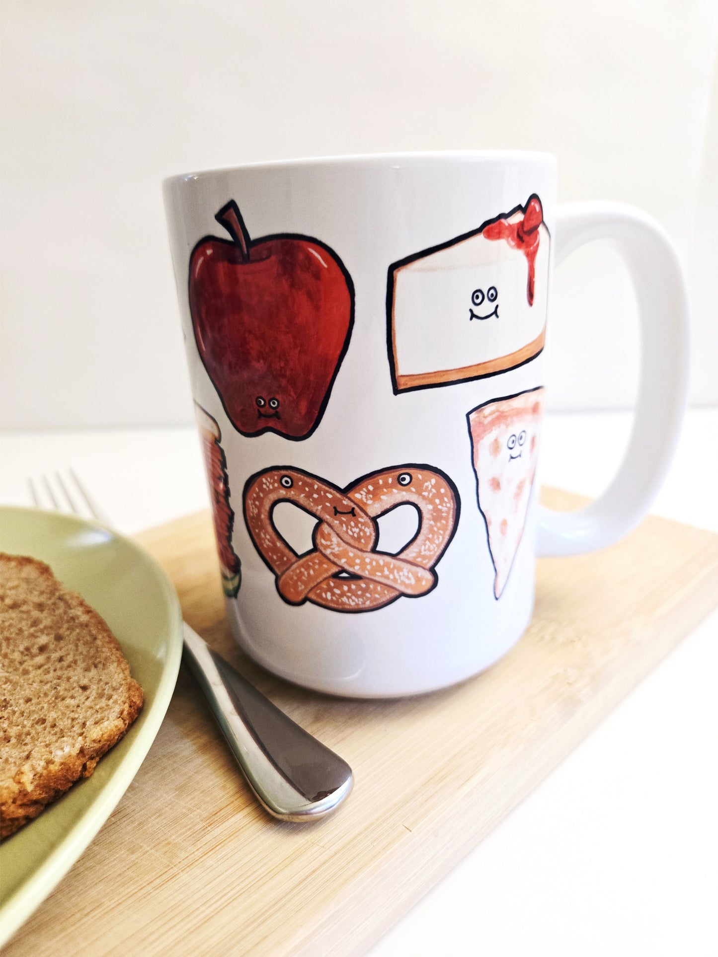White mug with NYC food-themed illustrations on a wooden surface with a plate and fork.